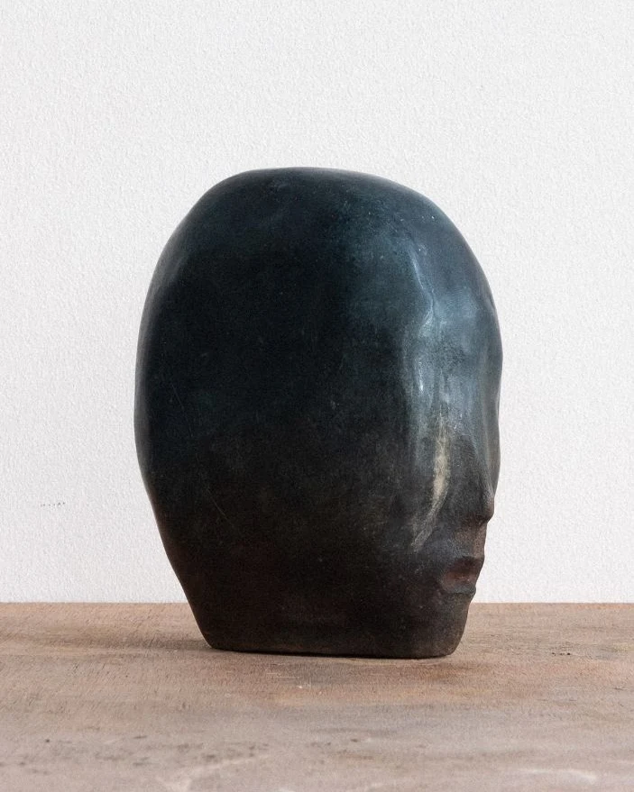 Blue Entity Study (2) by Arran Gregory, a dark blue-green burnished clay head with faint features, in profile on a wooden table