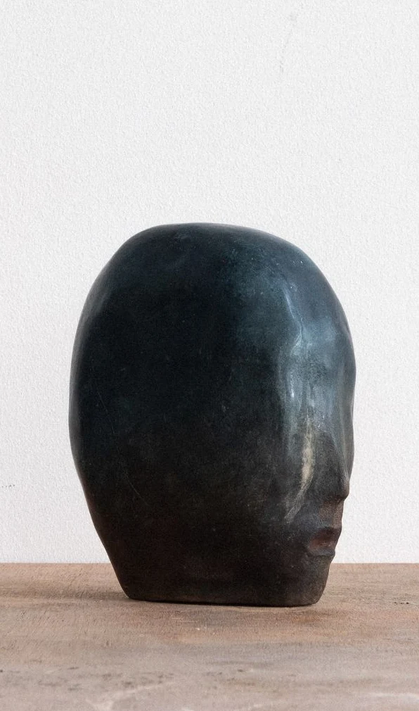 Blue Entity Study (2) by Arran Gregory, a dark blue-green burnished clay head with faint features, in profile on a wooden table