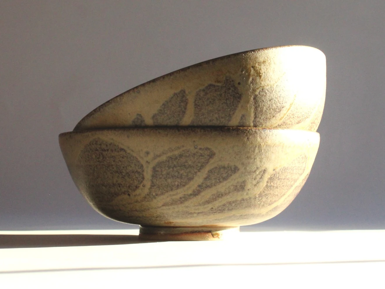 Large Bowl by James Galbraith Kirkpatrick, two stacked stoneware bowls with a speckled cream glaze and grey leaf-like marks, in low sunlight